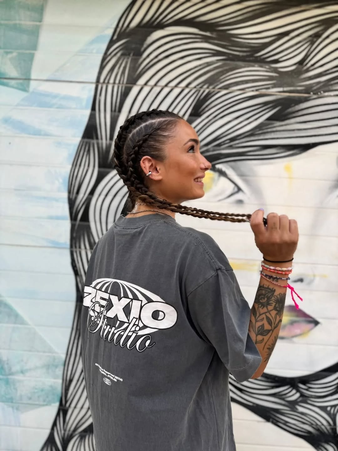 Se ve una mujer con trenzas y tuatuajes de flores llevando una camiseta de la marca de ropa Zexio Studio Gris
